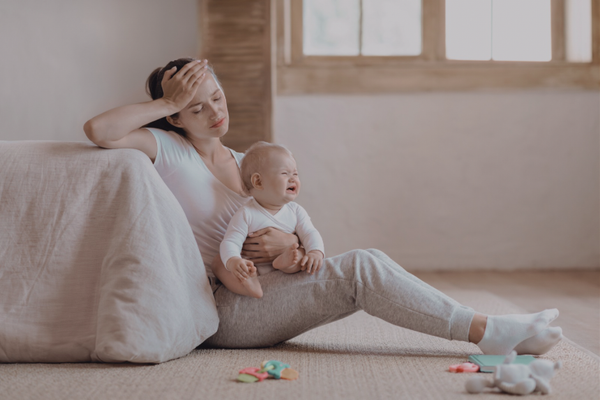 Overwhelmed mom sitting on the floor leaning on a bed with a baby secured in her lap
